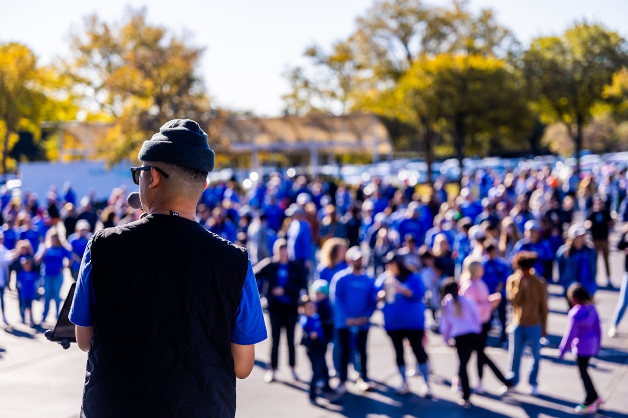 A speaker addresses a large crowd gathered outdoors, signifying a community event with a lively atmosphere.